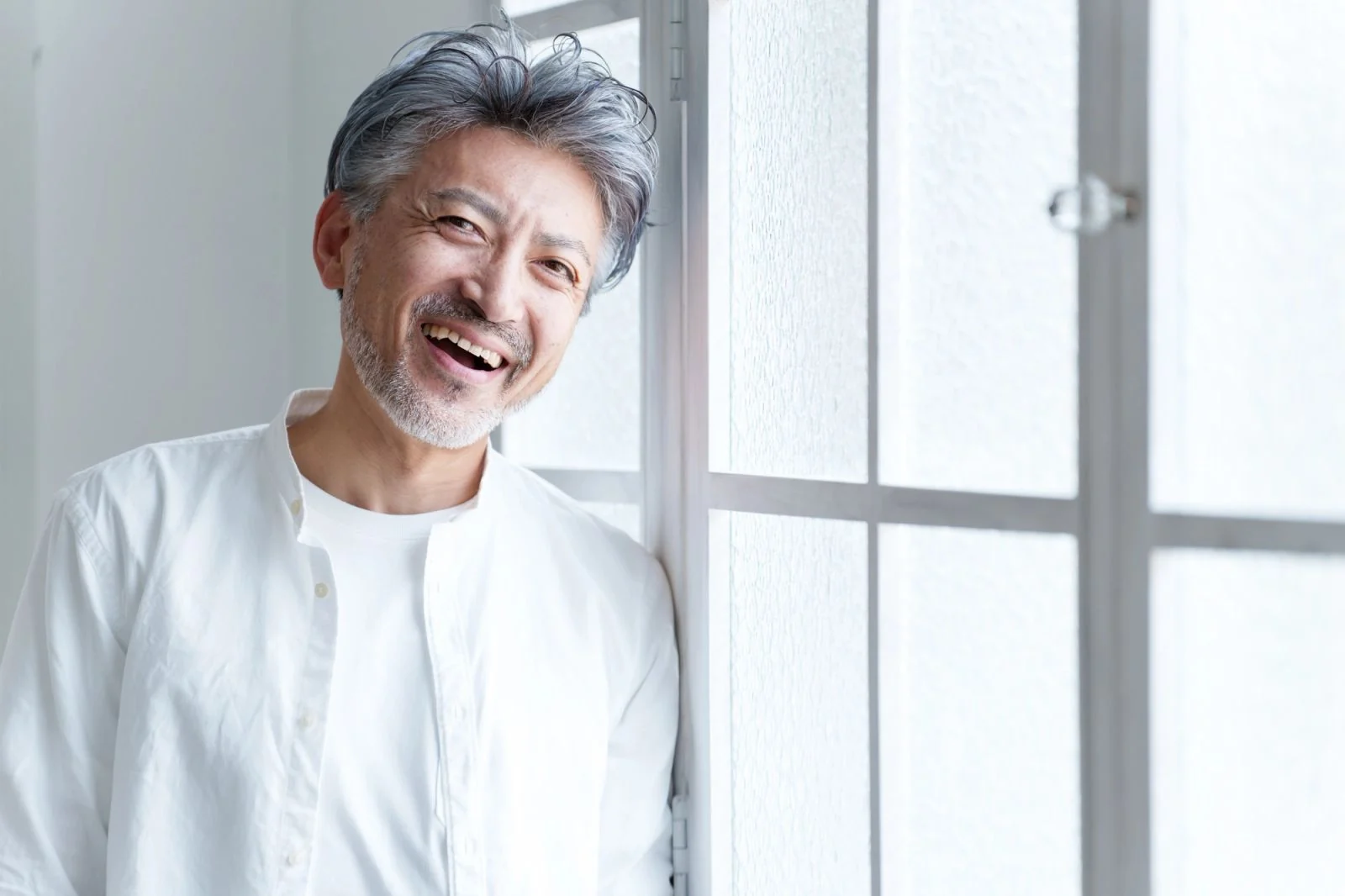 a man with gray hair and a beard is leaning against a window and smiling .