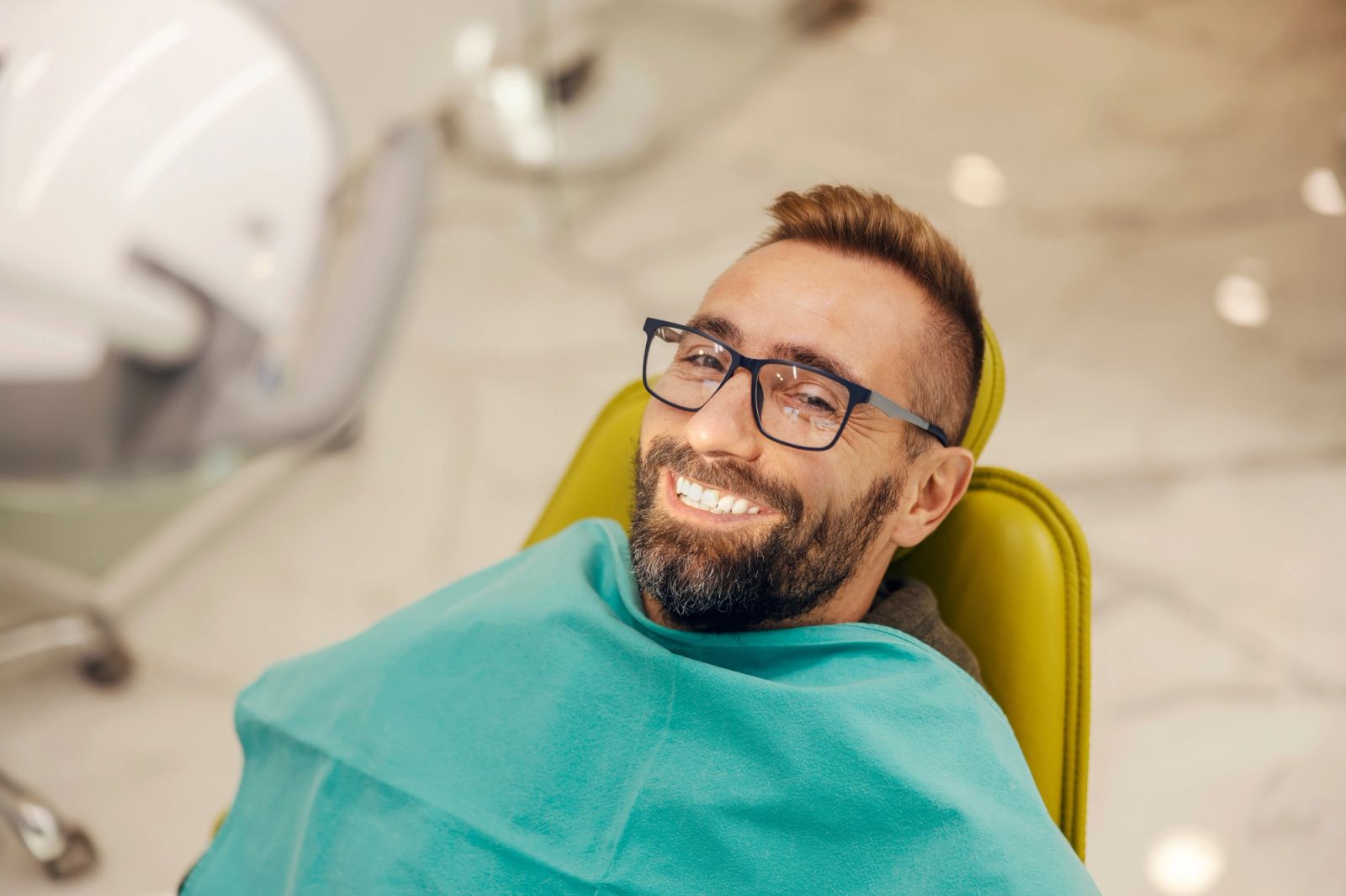 A smiling man with glasses and a beard in a dental chair.