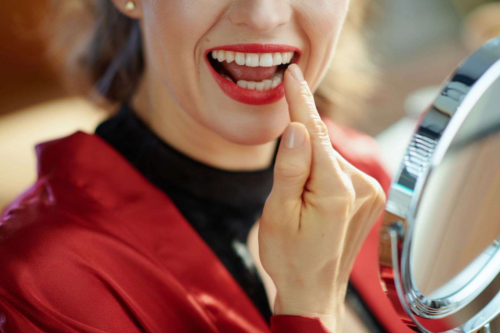 A woman in red lipstick and a red robe touching her teeth while looking into a mirror.