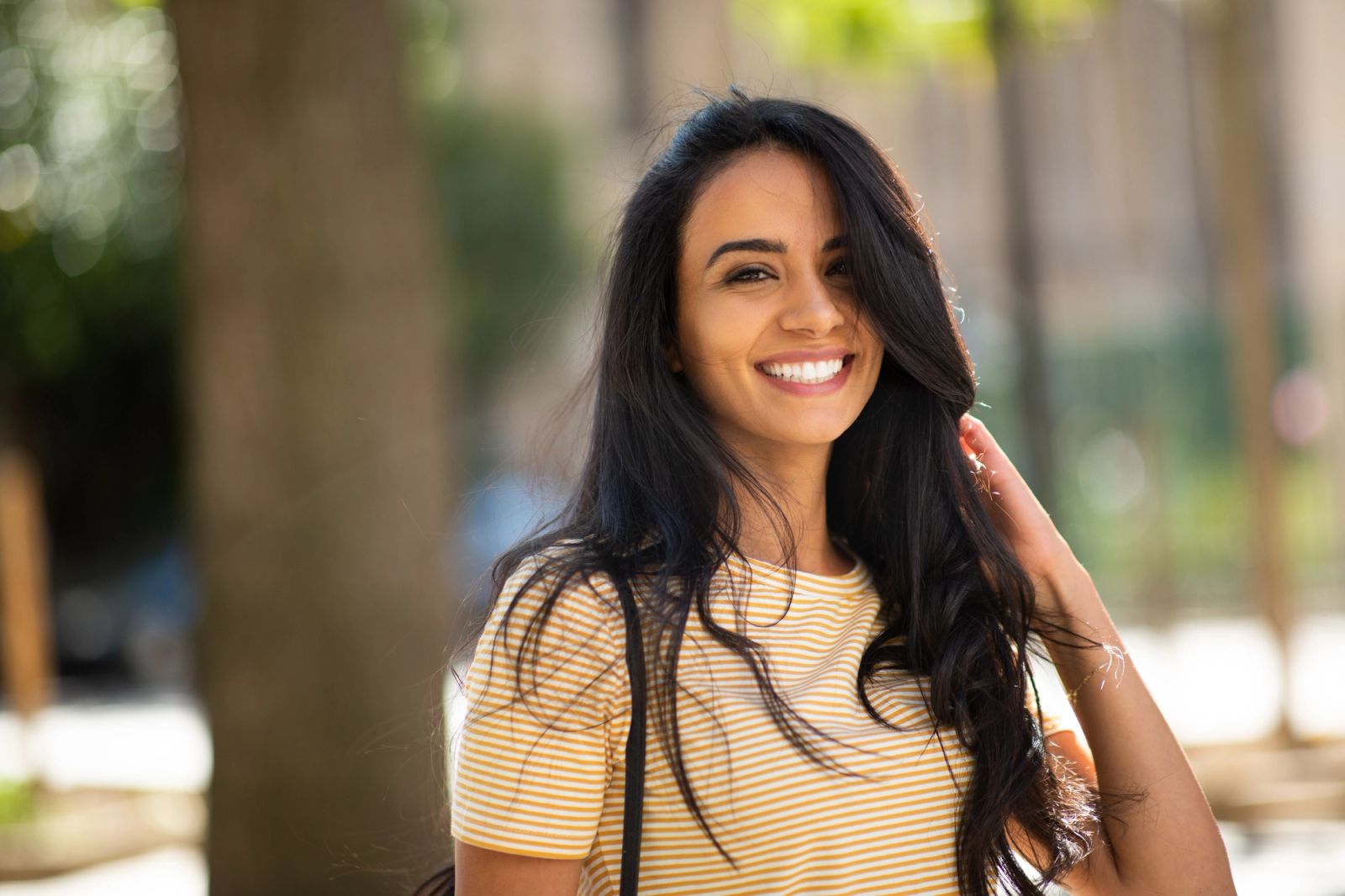 A smiling woman with long dark hair wears a yellow striped shirt outdoors.